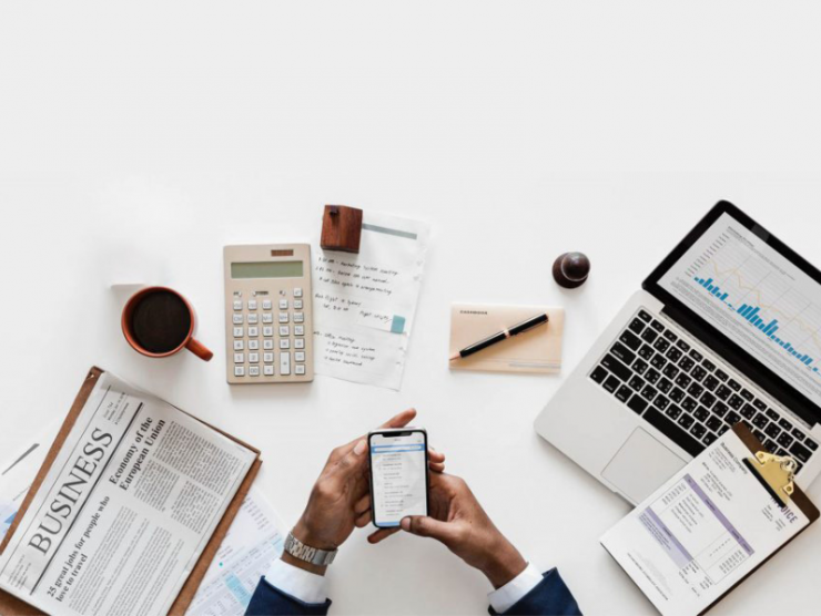 Overheard photo of a work desk with newspaper, calculator, laptop and a person's hands visible holding a phone while checking email.