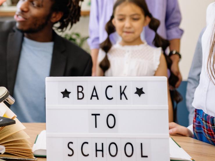 Several children and a teacher looking at a text book and example items, with a sign in the foreground reading "Back to School"